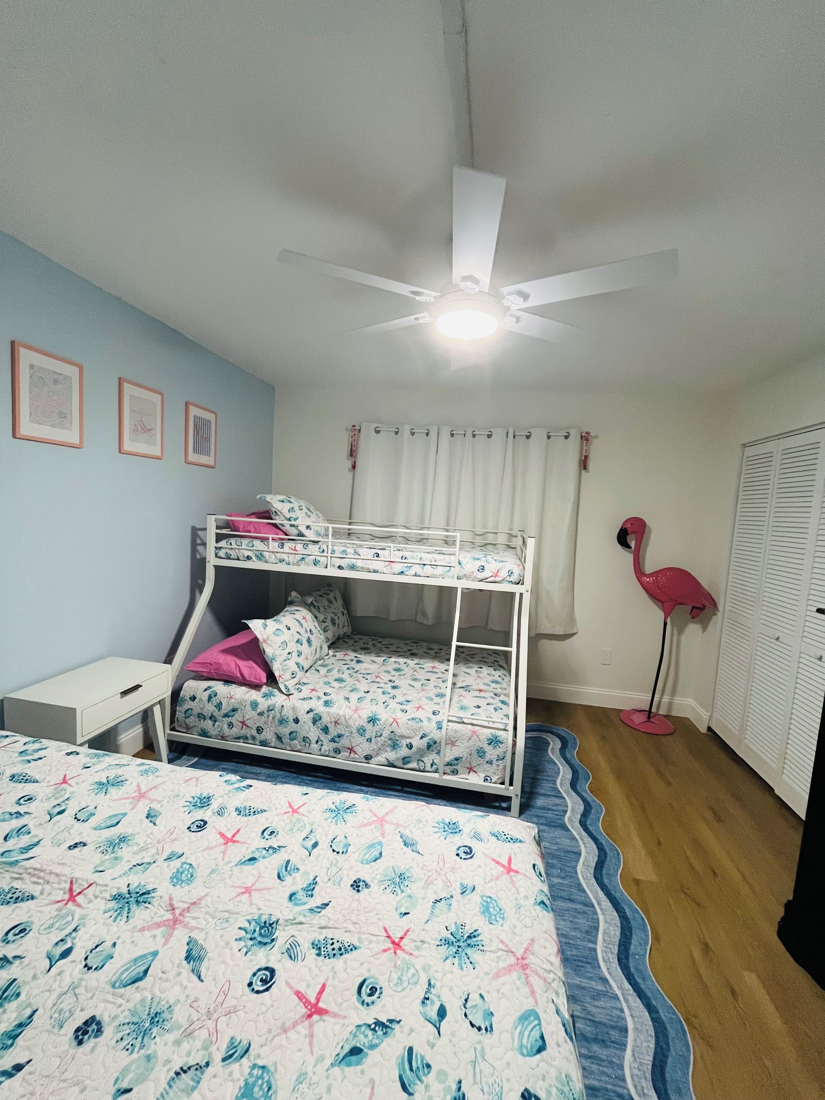 Ein Zimmer mit Etagenbetten, einem Schreibtisch, einer rosa Flamingo-Stehlampe und einem Deckenventilator.