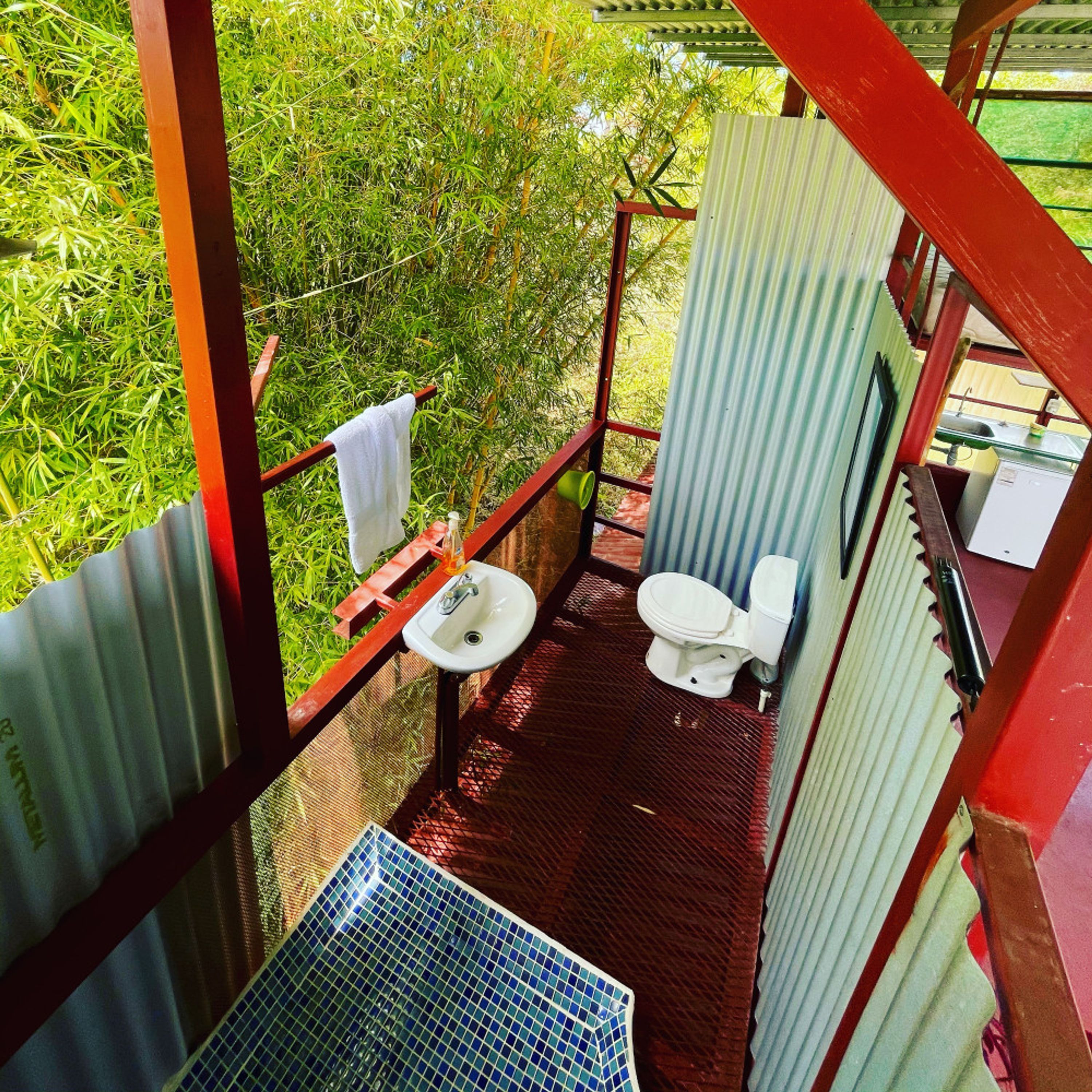 A small, enclosed outdoor bathroom with a toilet, sink, and a towel hanging on a rail.