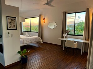 A hotel room with a bed, a desk, a chair, a potted plant, a wall-mounted mirror, and a view of the outdoors. - Villas Jacquelina (Quepos)