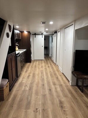 A narrow hallway with wooden flooring, white walls, and a kitchen area on the left with dark wooden cabinets and a white countertop.