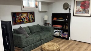 Un soggiorno con un divano verde, un pouf rotondo intrecciato, un mobile TV nero e un quadro appeso al muro.