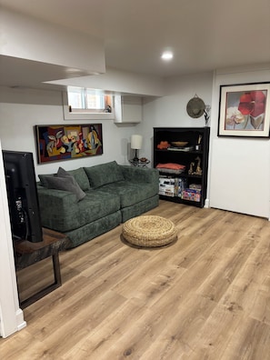 Uma sala de estar com um sofá verde, um banquinho redondo de fibra natural, um rack preto para eletrônicos e um quadro na parede.
