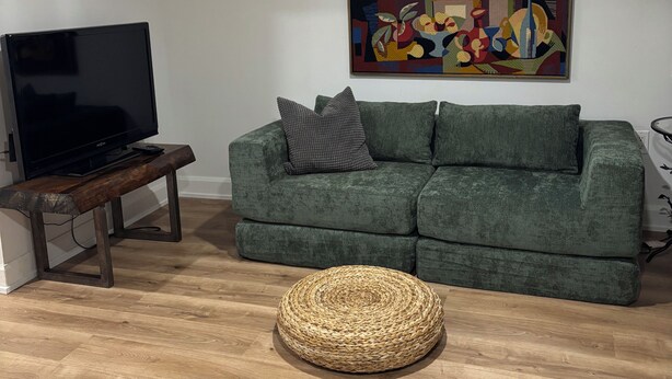 Un soggiorno con un divano verde, una televisione a schermo piatto su una panca in legno, un poggiapiedi rotondo intrecciato e un quadro astratto colorato appeso al muro.