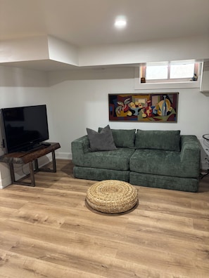 A living room with a green sofa, a flat-screen TV on a wooden stand, a woven round ottoman, and a colorful abstract painting on the wall.