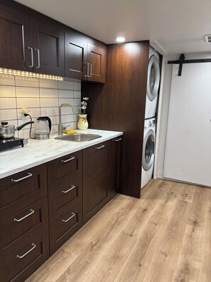 A kitchen with dark wooden cabinets, a stainless steel sink, a kettle, and a washing machine. - Cozy East York Suite | Private & Quiet | Close to Transit (Toronto)