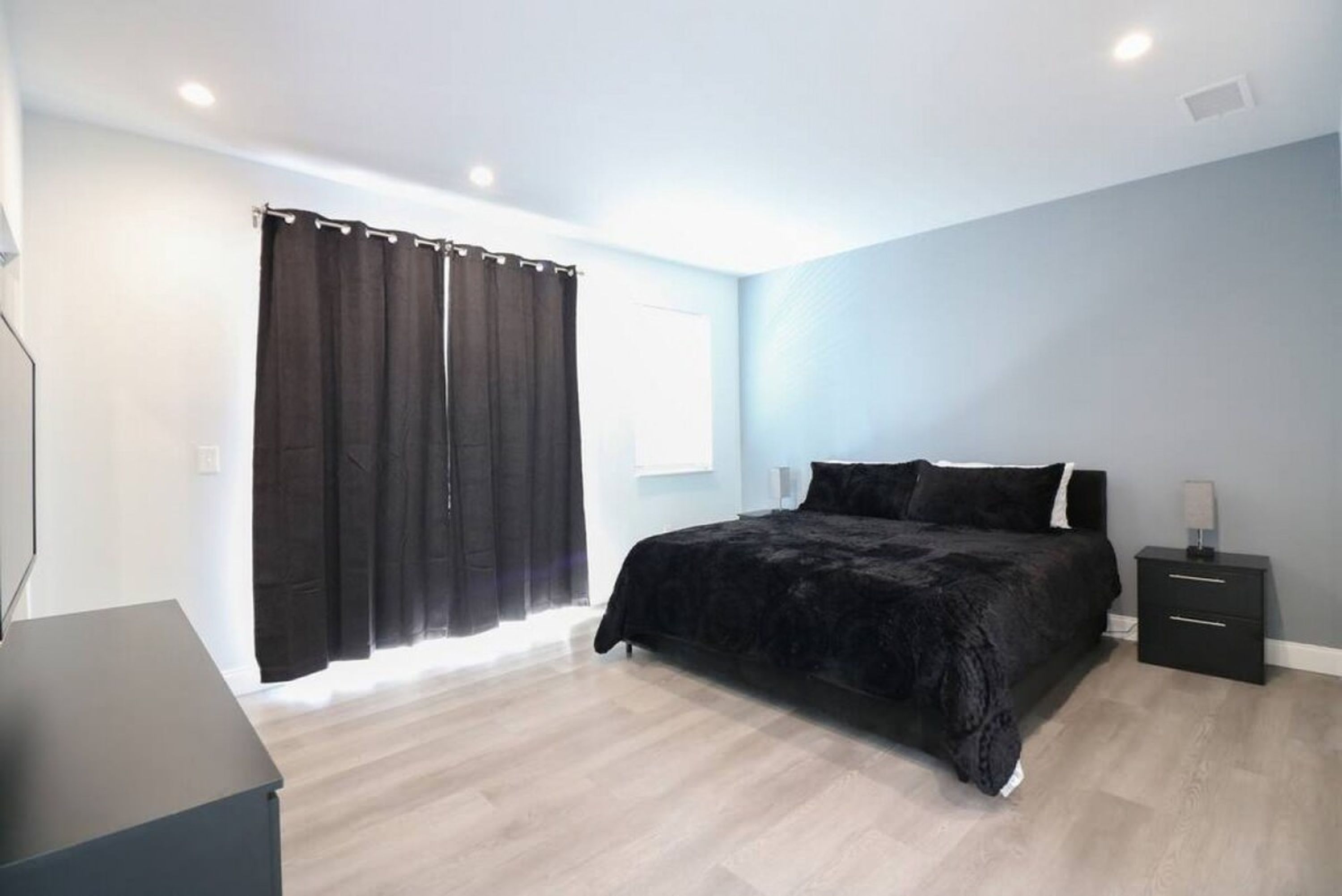 Ein Schlafzimmer mit Bett, Nachttisch, Kommode und einem Fenster mit Vorhängen.