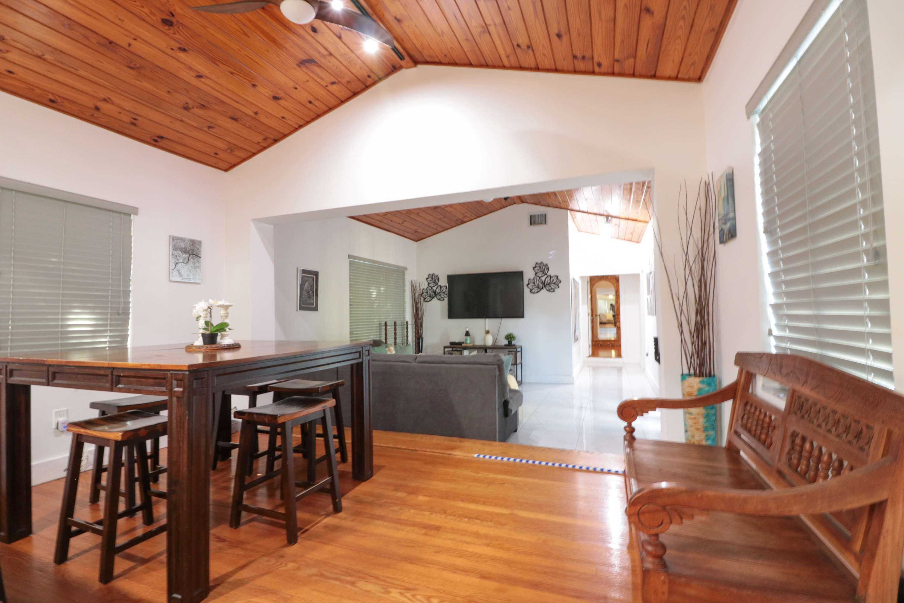 Uma sala de estar ampla com teto de madeira, uma mesa de jantar com banquetas, um sofá e uma televisão fixada na parede.