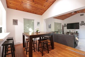 Un soggiorno moderno con soffitto in legno, un'area pranzo con tavolo e sedie, un divano grigio e una TV a schermo piatto montata a parete.