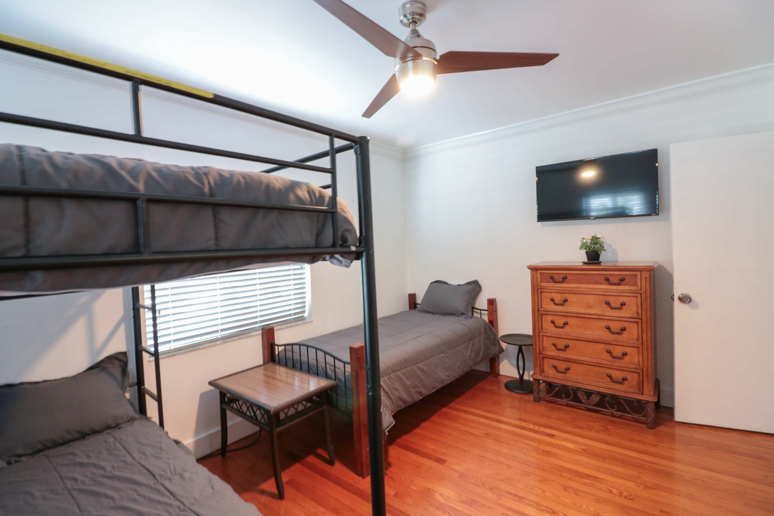 A room with a bunk bed, a single bed, a dresser, a television, and a ceiling fan.