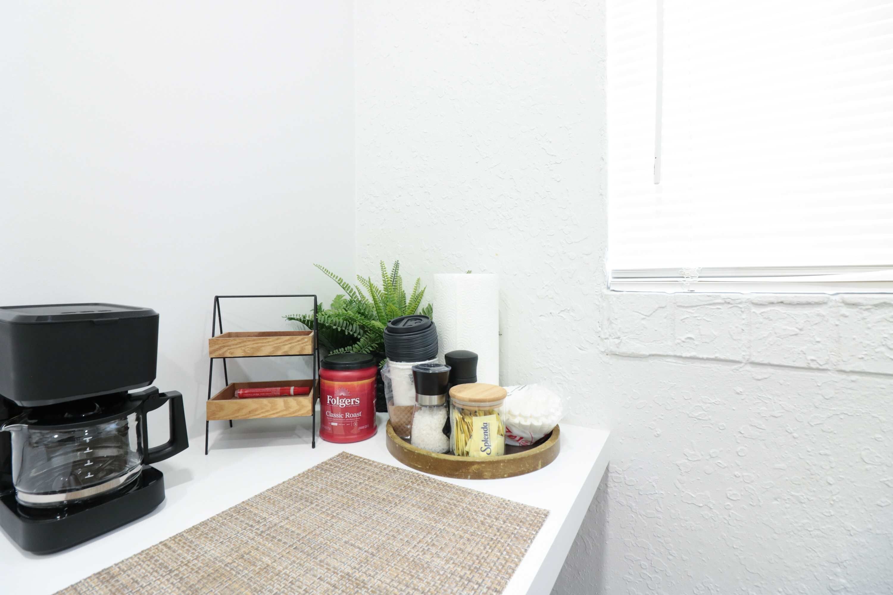 A kitchen counter with a coffee maker, a wooden rack with a plant, and various kitchen items like a jar of coffee, a container of sugar, and a spice jar.