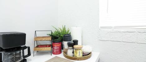 A kitchen counter with a coffee maker, a wooden rack with a plant, and various kitchen items like a jar of coffee, a container of sugar, and a spice jar.