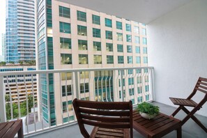 A balcony with a view of a modern building with multiple windows. There are two wooden chairs and a small table with a plant. - Luxury Brickell Condo 1 BR Resort Amenities (Miami)