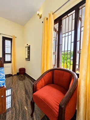 A room with a large window, a chair, a bed with a blue patterned bedspread, and a small round ottoman.