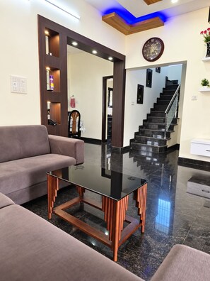 A modern living room with a glass coffee table, a sofa, a staircase, and a clock on the wall.
