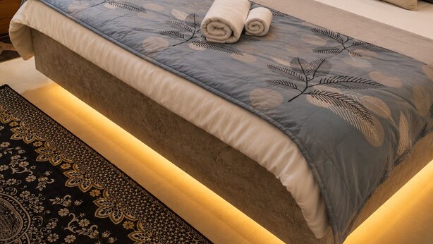 Een netjes opgemaakt bed met een bedsprei met patroon, een hoofdbord en een nachtkastje met een lamp.
