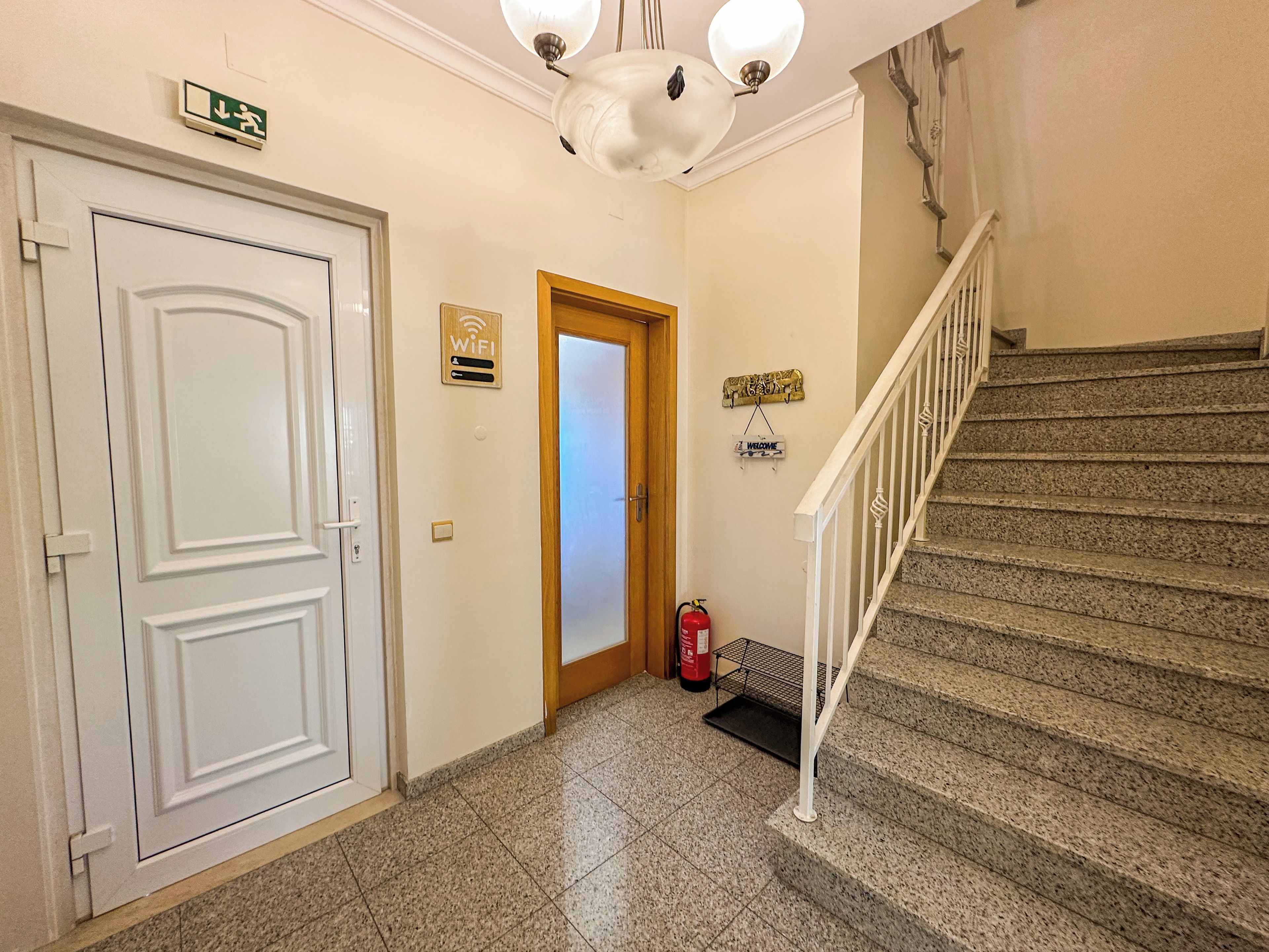 A hallway with a white door, a wooden door, a sign indicating WiFi, a welcome sign, a fire extinguisher, and a staircase.