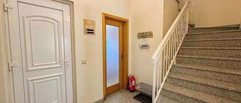 A hallway with a white door, a wooden door, a sign indicating WiFi, a welcome sign, a fire extinguisher, and a staircase.