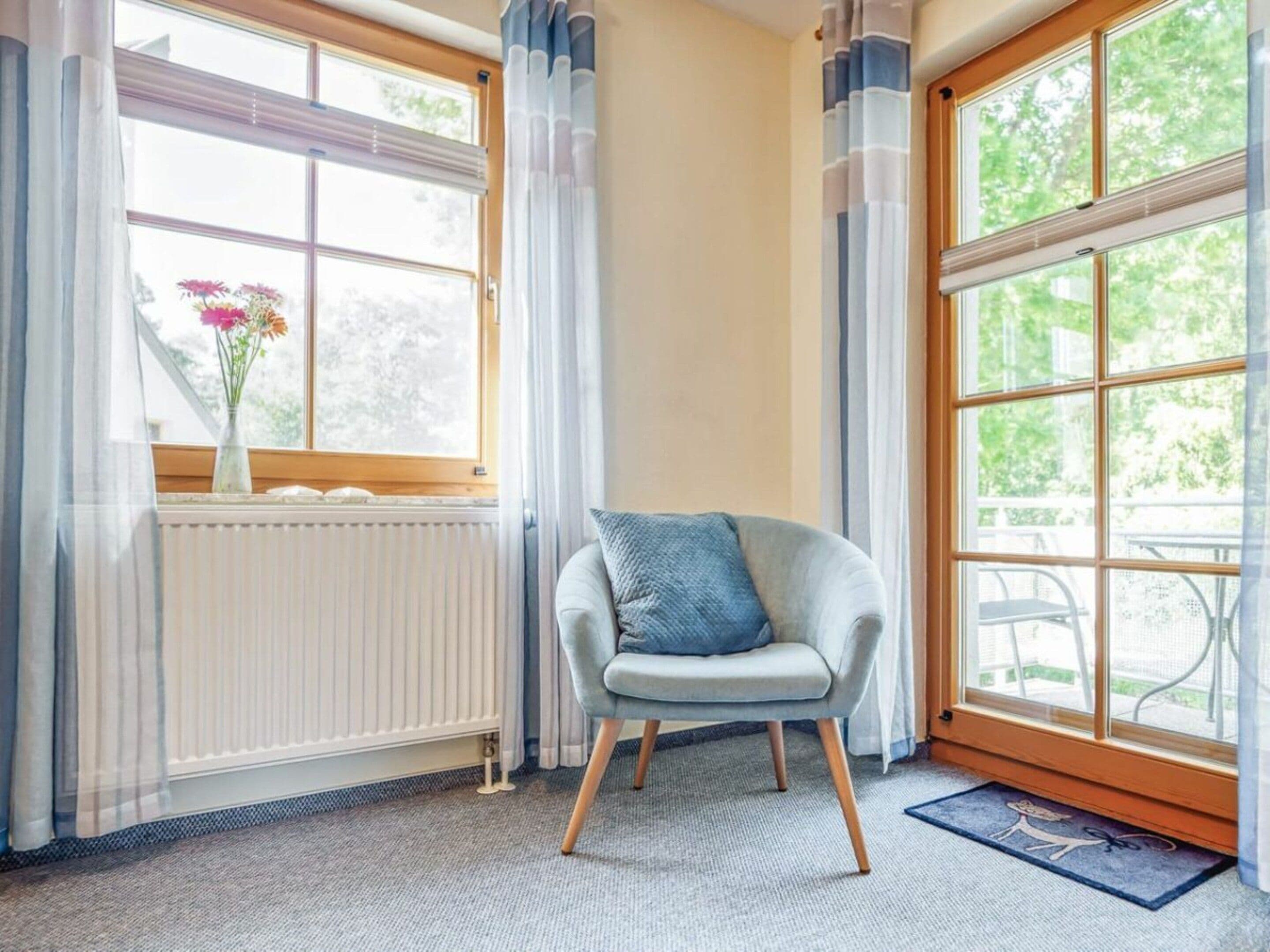 Ein Eckzimmer mit Heizkörper, einem Sessel mit Kissen und großen Fenstern mit Vorhängen.
