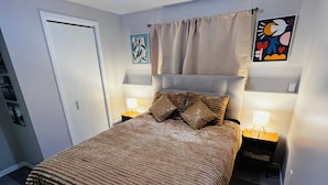 A bedroom with a bed, two bedside tables, a ceiling light fixture, a door, and a wall with a framed picture. - Cozy 1 Bdr Apartment (Mount Pearl)