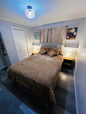 Un dormitorio con una cama, dos mesitas de noche, una lámpara del techo, una puerta y una pared con un cuadro enmarcado.