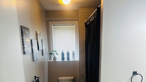 A compact bathroom with a toilet, a small window, a shower curtain, and a towel rack. - Cozy 1 Bdr Apartment (Mount Pearl)