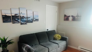 A living room with a grey sofa, a small table with a plant, and wall art depicting coastal scenes. - Cozy 1 Bdr Apartment (Mount Pearl)