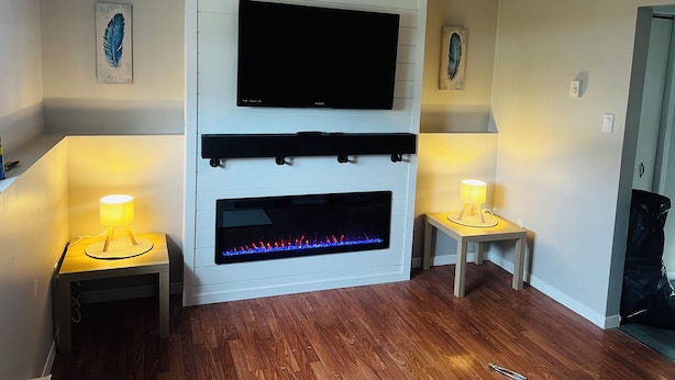 A living room with a flat-screen TV mounted above a fireplace, flanked by two small tables with lamps. - Cozy 1 Bdr Apartment (Mount Pearl)