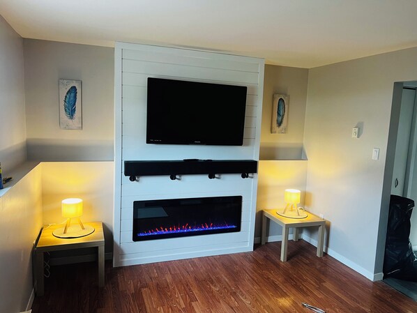 Un salón con una televisión de pantalla plana sobre una chimenea, flanqueada por dos mesitas con lámparas.