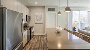 Una cocina moderna con electrodomésticos de acero inoxidable, suelo de madera, una isla central, iluminación colgante y vistas al exterior.