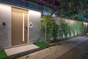 Un edificio moderno con una entrada bien iluminada, un pequeño espacio exterior con césped artificial y una pared cubierta de plantas trepadoras.