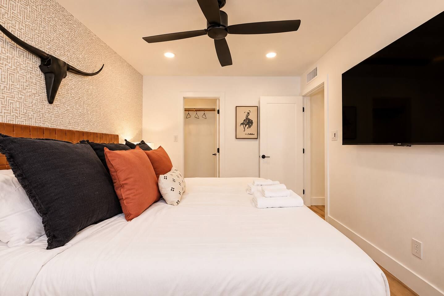 Una habitación de hotel con una cama, un ventilador de techo, un televisor montado y una puerta que da a otra habitación.