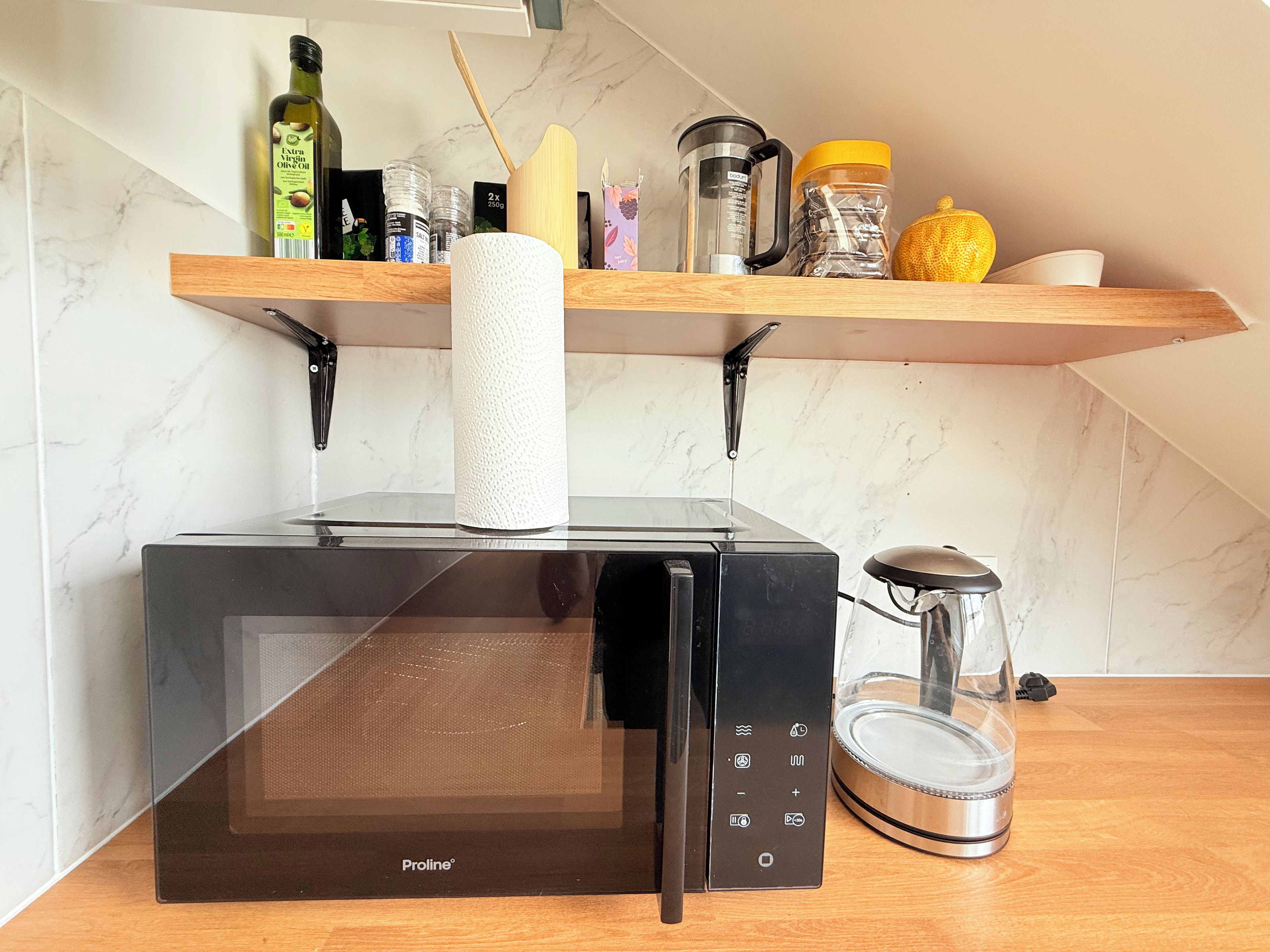 Una cocina con un microondas negro, una cafetera de vidrio y una repisa de madera con varios utensilios de cocina.