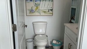 Casa de banho com sanita, um caixote do lixo, uma cesta e um quadro com uma cena de praia.