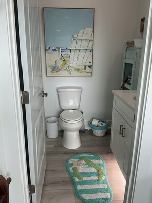 A bathroom with a toilet, a trash bin, a basket, and a painting of a beach scene. - Beautiful Beach Home on the West End of Galveston with Beach across the street (Galveston)