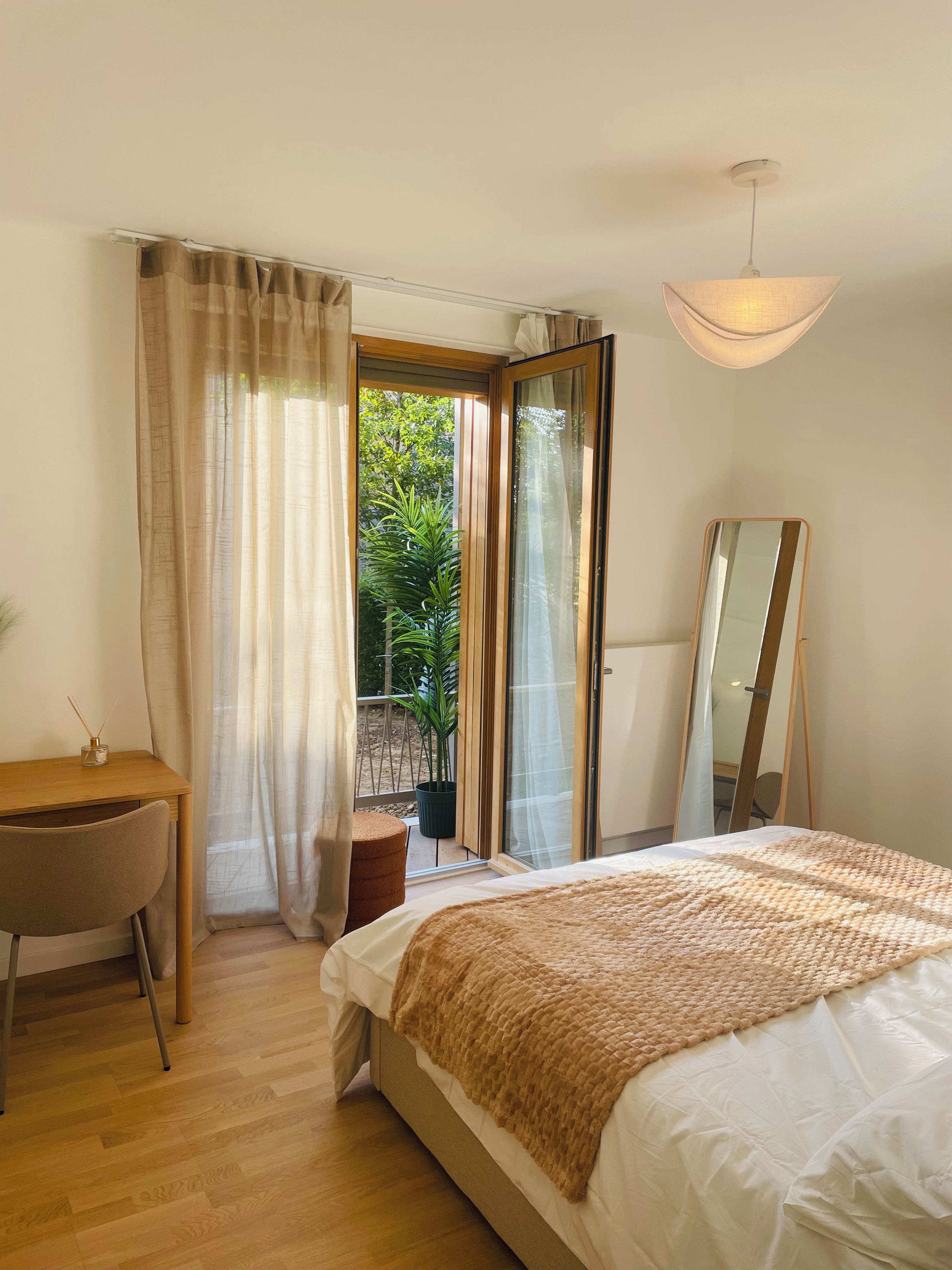 A bedroom with a bed, a desk, a chair, a mirror, and a view of greenery outside.