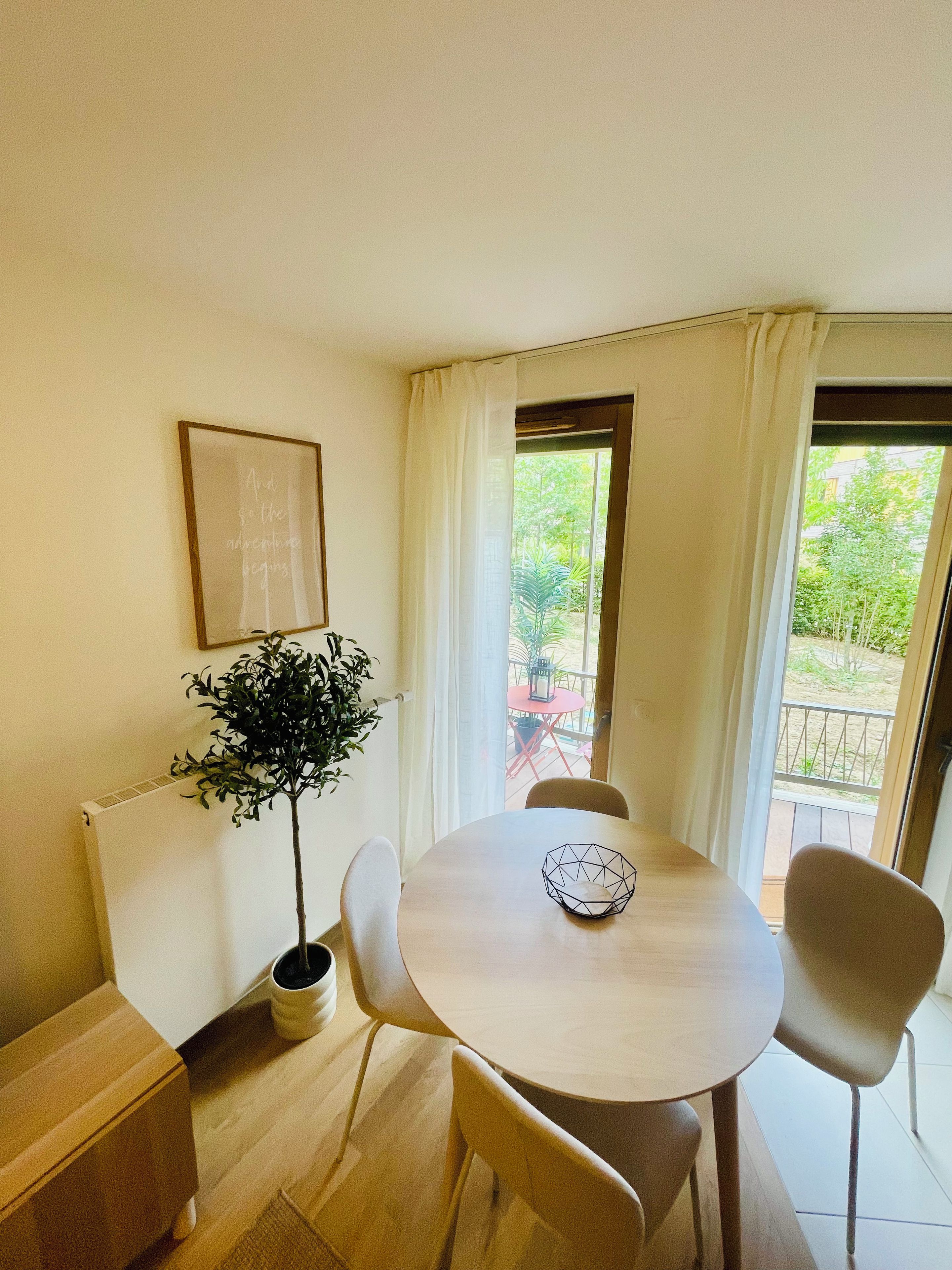 Un coin repas avec une table ronde, quatre chaises, une plante en pot, un tableau encadré et une vue sur l’extérieur par une fenêtre.