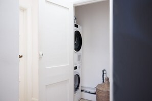Une buanderie équipée d’un lave-linge et d’un sèche-linge superposés, d’un panier en osier et d’une porte blanche.