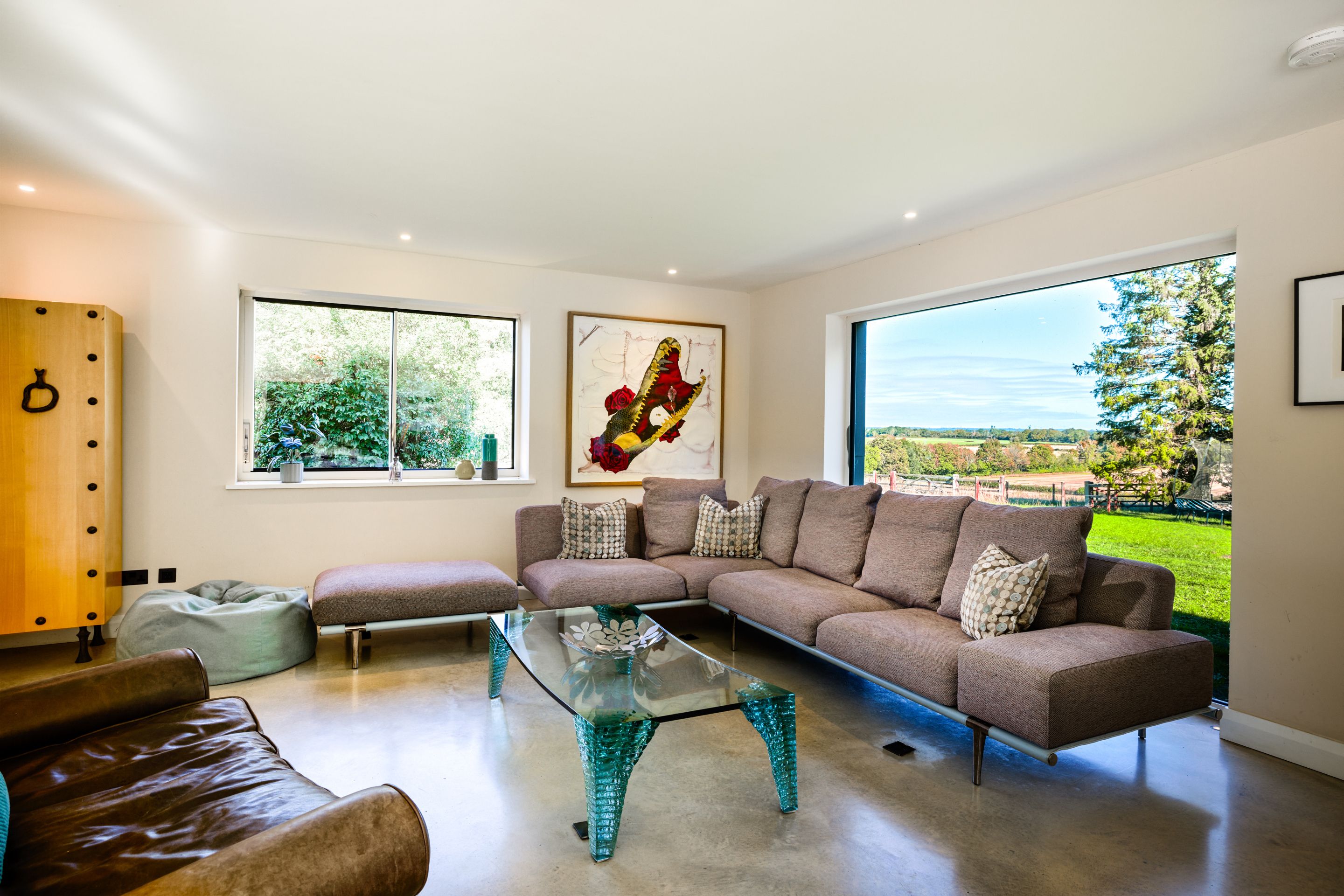 A modern living room with a sectional sofa, a glass coffee table, and a view of a park through large windows.
