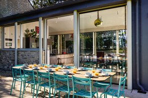 Un espace de restauration en plein air avec des tables et des chaises, un mur en pierre et une grande fenêtre donnant sur un jardin.