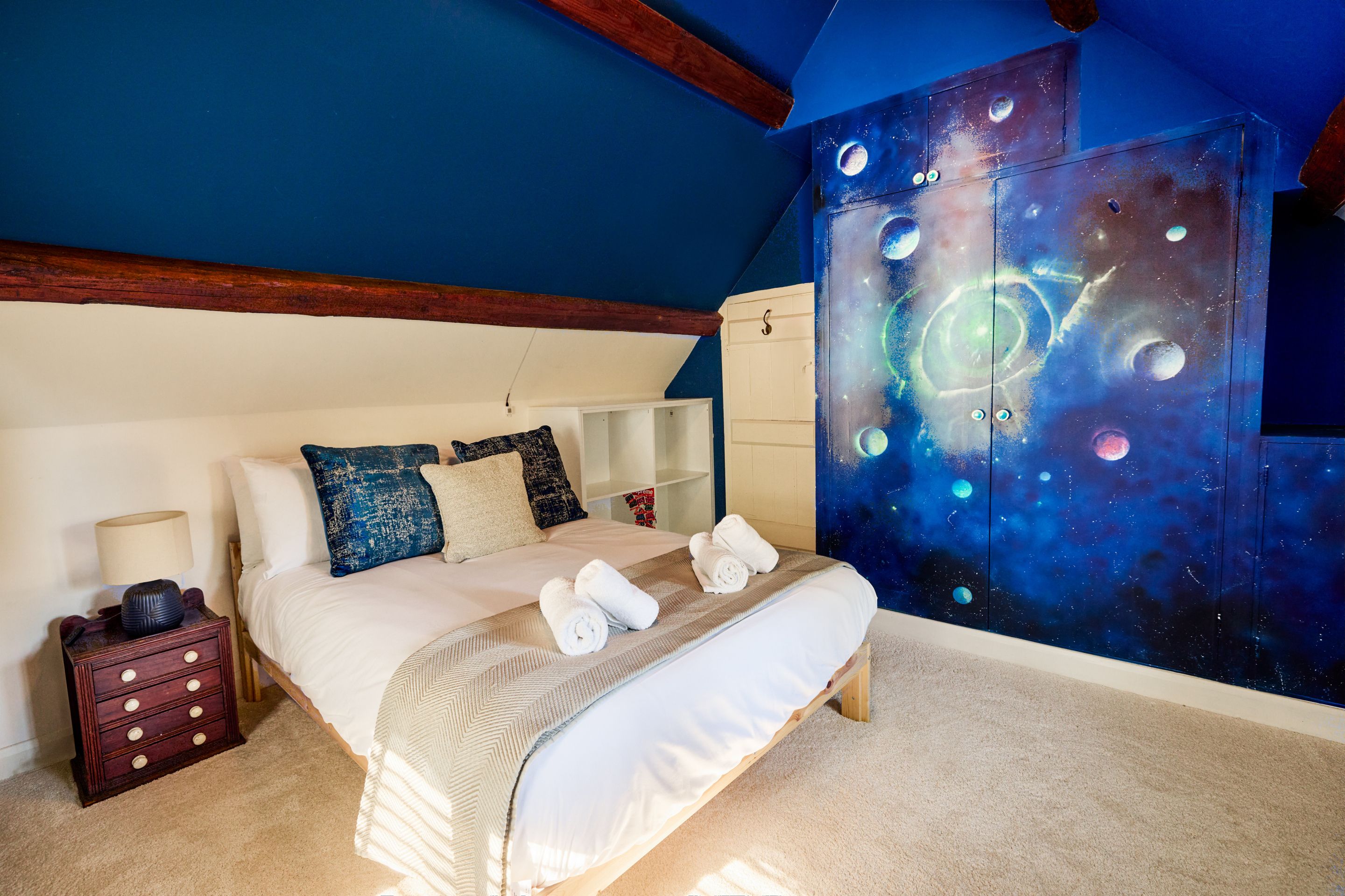 Een slaapkamer met een bed, nachtkastje, kapstok en een muur met een ruimtethema-muurschildering.