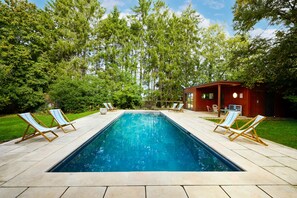 Une piscine rectangulaire à l’eau d’un bleu limpide, entourée d’une terrasse carrelée. De chaque côté de la piscine, on trouve deux chaises longues en bois avec des coussins à rayures.