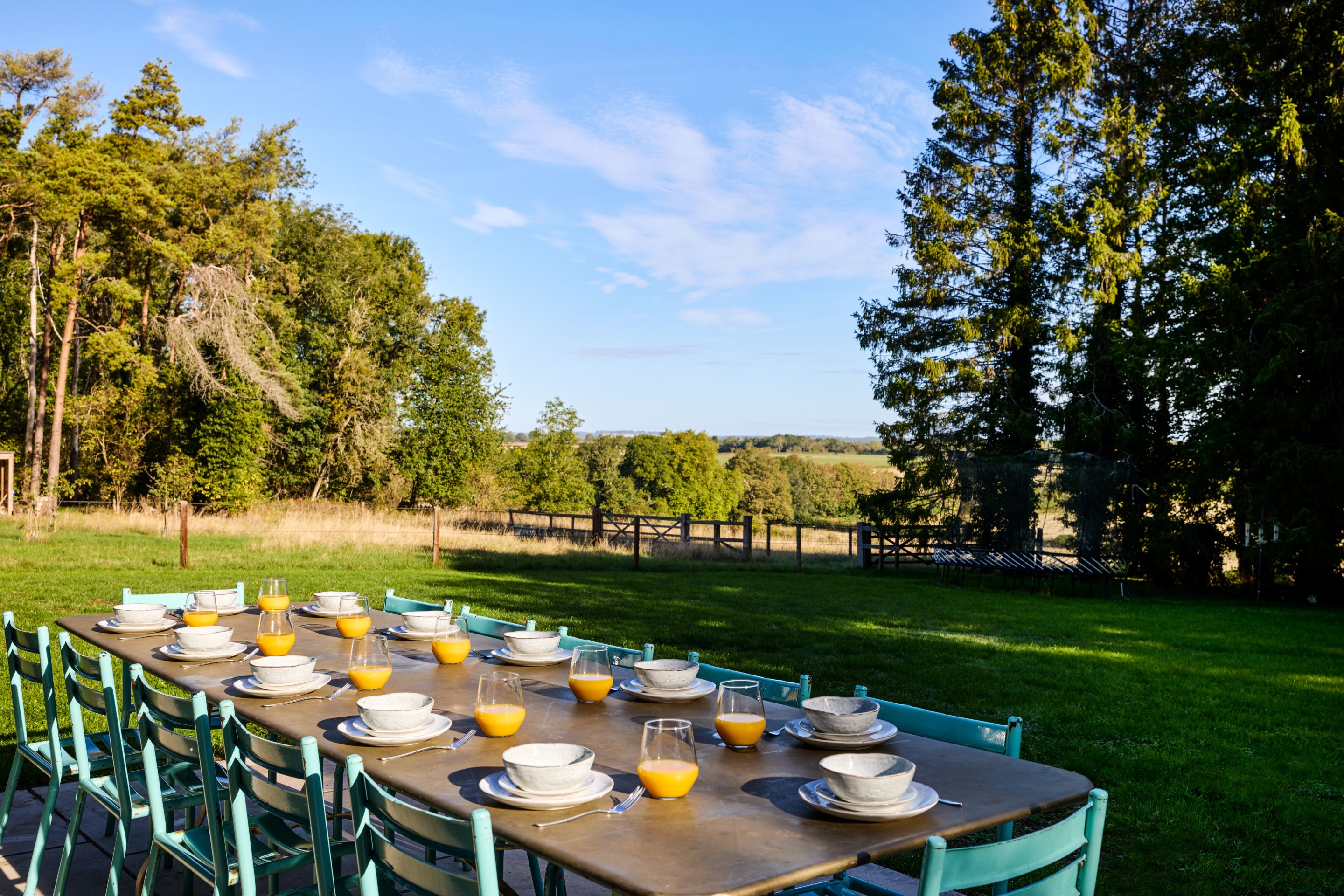 An outdoor dining area with a long table set for breakfast, surrounded by chairs, with a view of trees and a fence in the background.