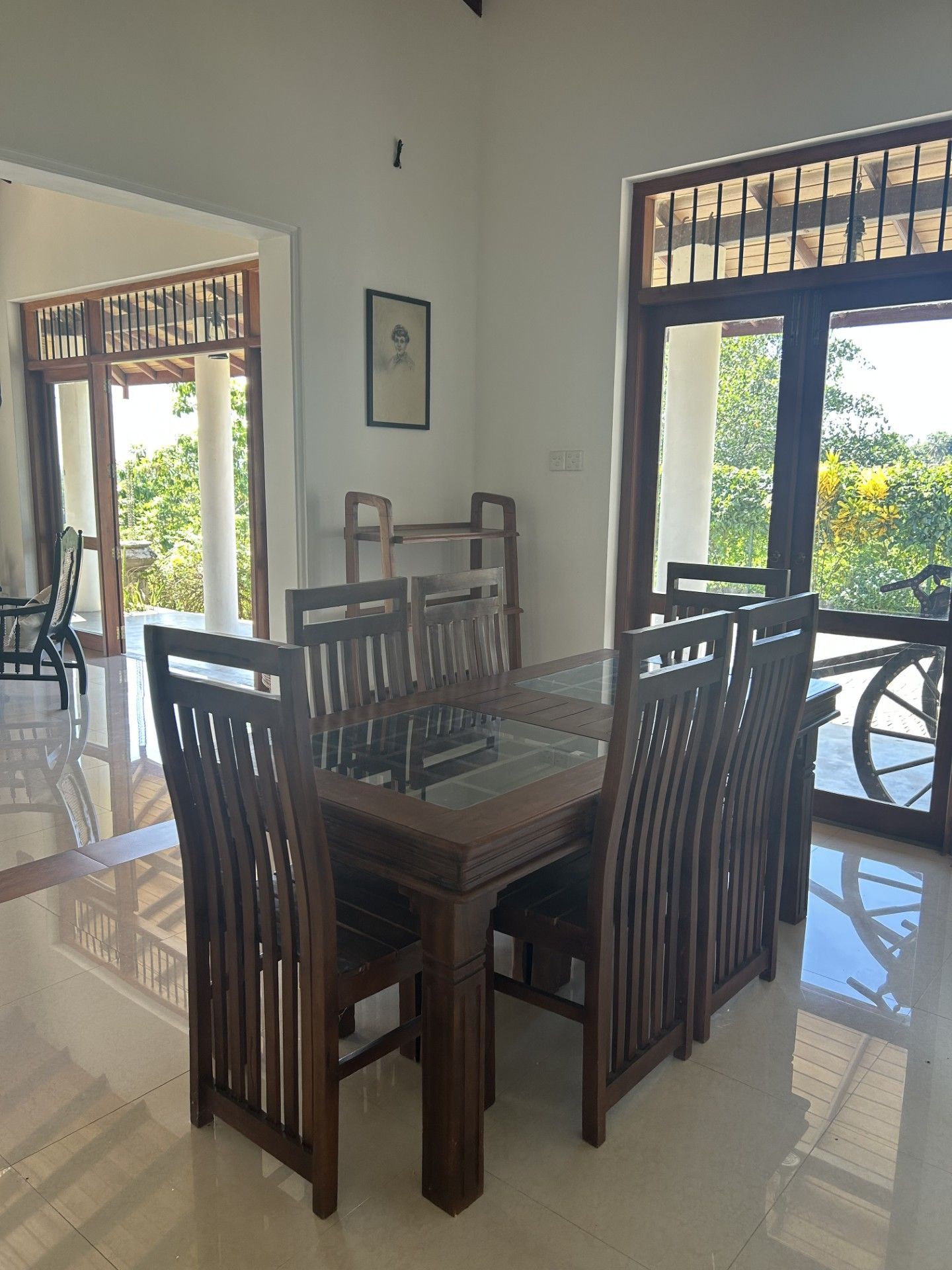 A dining area with a wooden table and chairs, a framed picture on the wall, and large windows with a view of greenery.