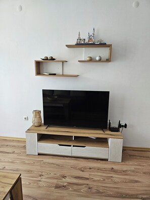 A modern living room with a flat-screen TV on a wooden stand, two wall-mounted shelves with decorative items, and a woven basket on the floor. - Chateau apartment (Aheloy)