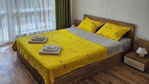 A neatly made bed with a yellow bedspread and pillows, a wooden headboard, a bedside table with a lamp, and a window with sheer curtains. - Chateau apartment (Aheloy)