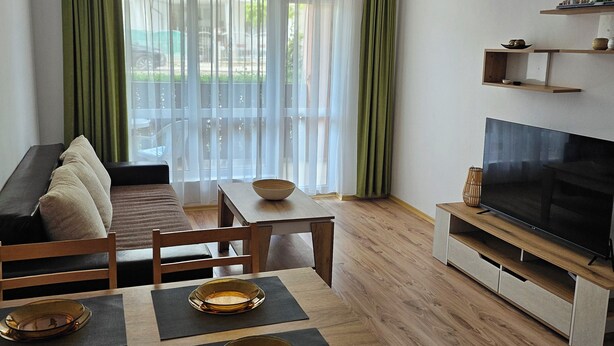 A modern living room with a sofa, dining table, chairs, a flat-screen TV, and a balcony with curtains. - Chateau apartment (Aheloy)