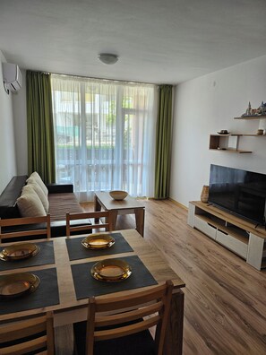 A modern living room with a sofa, dining table, chairs, a TV, and a balcony with curtains. - Chateau apartment (Aheloy)