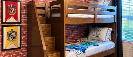 Ein Etagenbett mit Holzlade und Schubladen, eine Ziegelwand mit gerahmten Wappen, und ein Fenster mit Vorhängen.