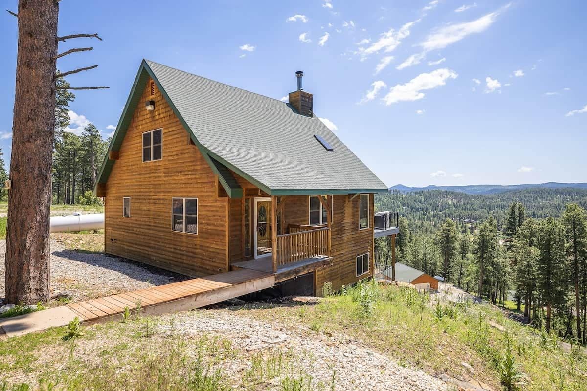 A log cabin with a green roof, a porch, and a view of the surrounding forest.