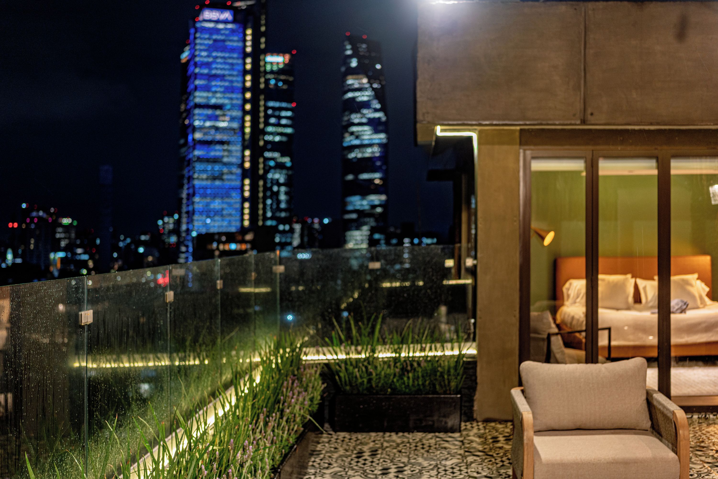 Una habitación de hotel moderna con vistas a la ciudad, separada por un panel de vidrio y con un balcón lleno de plantas.